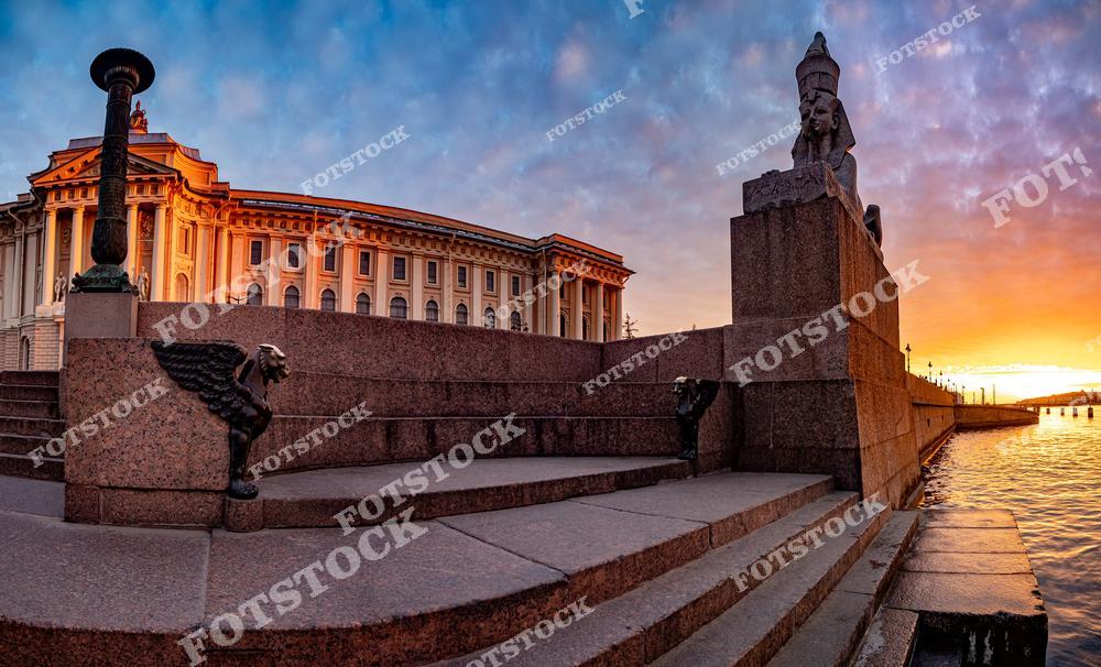 На фотографии изображено здание Академии художеств, освещенное мягкими лучами закатного солнца. На переднем плане видны знаменитые гранитные лестницы с фигурами грифонов и египетских сфинксов, украшающих Университетскую набережную. Яркий закат окрашивает небо и воды Невы в теплые тона, создавая атмосферу гармонии и величия. Это место является символом искусства и истории Петербурга.
